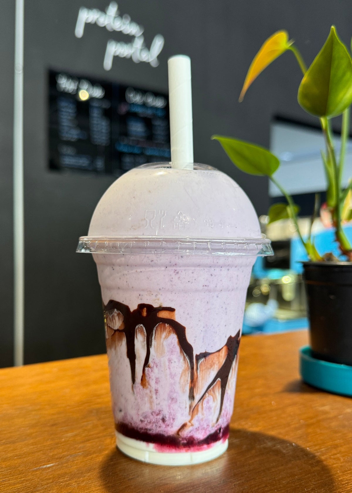 A purple iced coffee drink in a clear plastic cup with a white lid and straw, decorated with chocolate drizzle, sitting on a wooden table in a cafe.